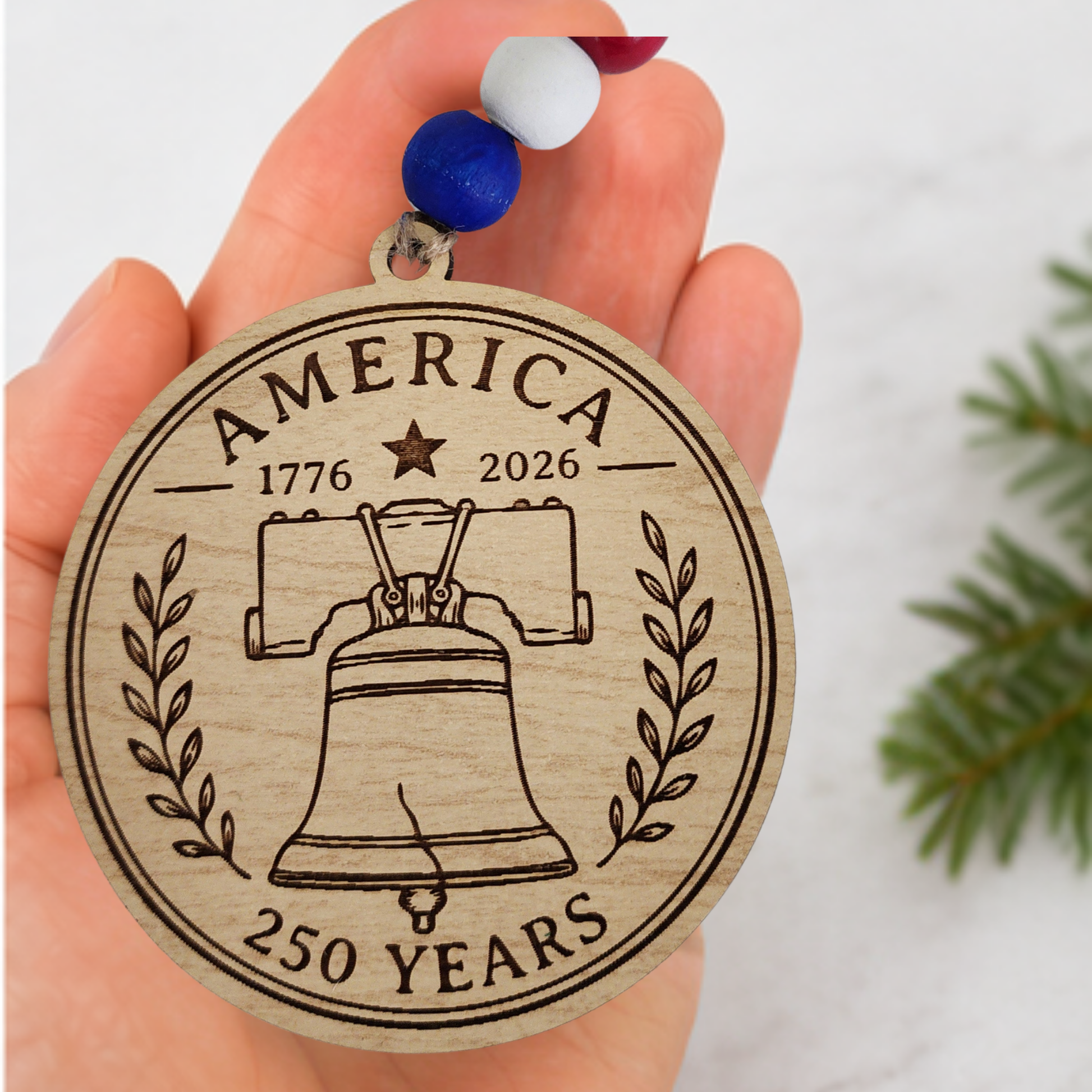 Handcrafted wooden America 250 ornament featuring Liberty Bell. Laser engraved patriotic keepsake with red, white, and blue bead accents celebrating America's 250th anniversary.